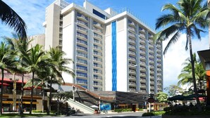 A multi-story hotel with a prominent blue stripe, palm trees, and a covered outdoor area. - HGVC Hokulani Waikiki Luxury, 1 BR Suites, Rooftop Pool/Spa, Close to Ft Derussy (Honolulu)