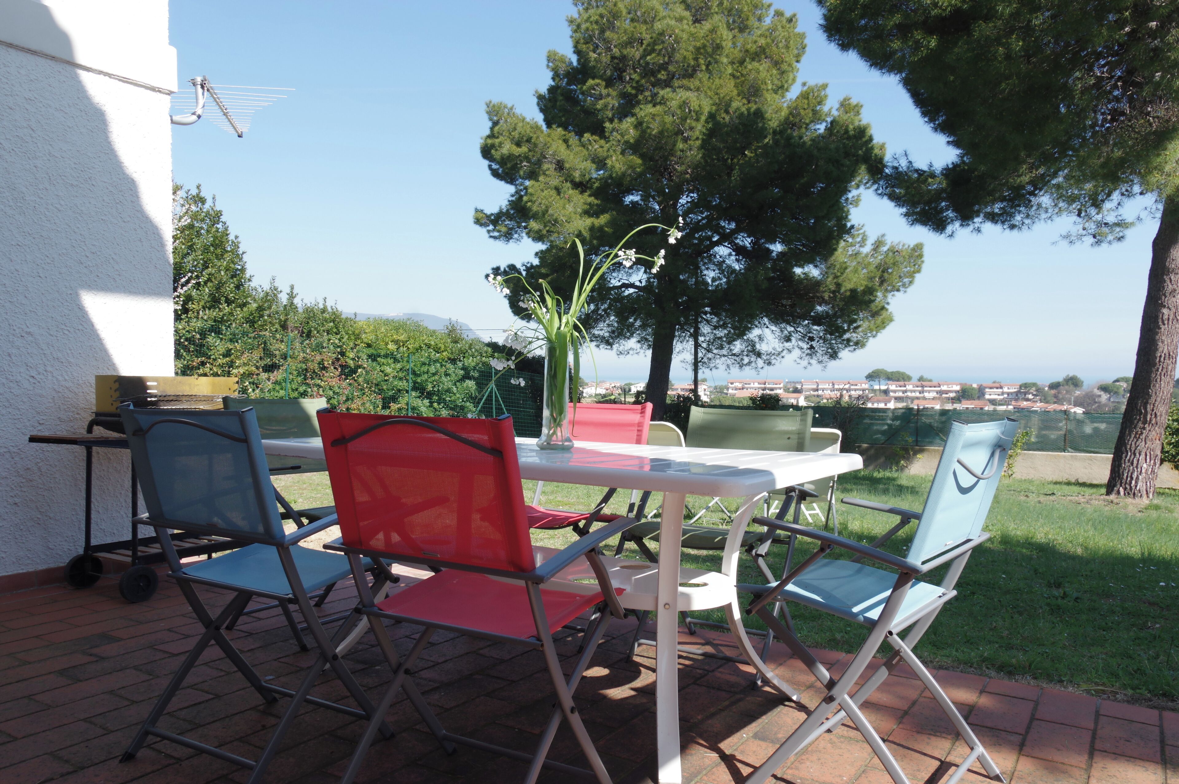Une terrasse extérieure avec une table blanche et quatre chaises, dont une rouge et trois bleues, offrant une vue panoramique sur un paysage verdoyant avec des arbres et des collines au loin.