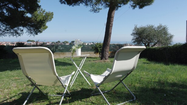 Two white lawn chairs facing each other with a small table between them, set on a grassy area with a tree and a view of a distant landscape.