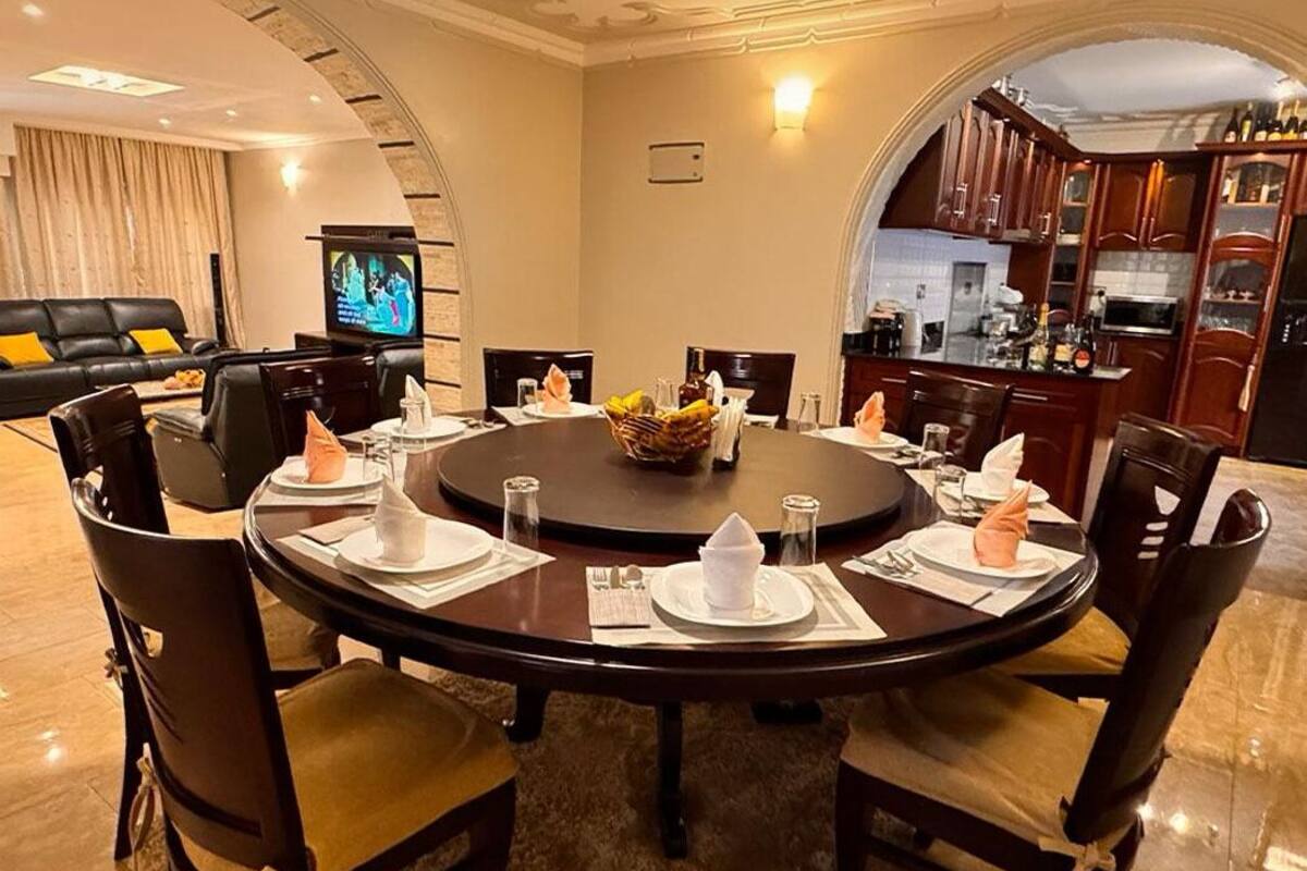 A round dining table set for six with dark wood chairs, a central fruit basket, and a television in the background.