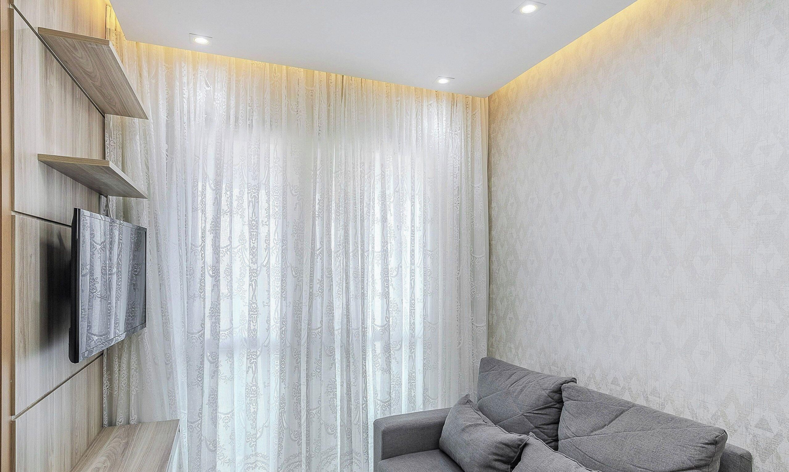 A modern living room with a flat-screen TV mounted on the wall, a grey sofa with cushions, and a large window with sheer curtains.