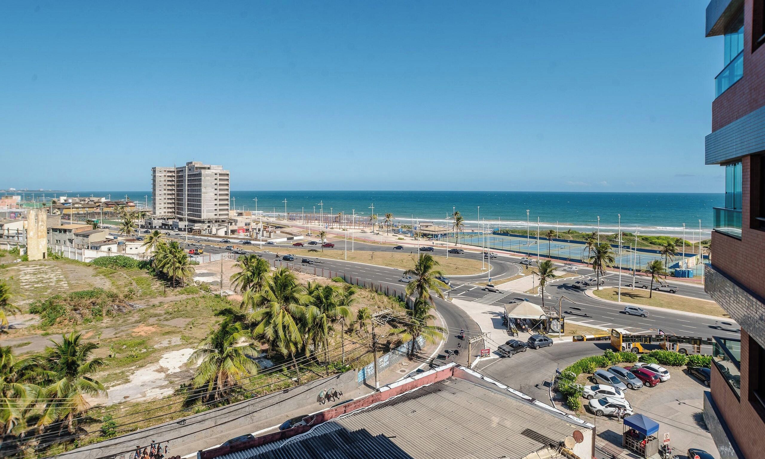 A coastal cityscape with a prominent high-rise building, a wide road with vehicles, and a beach in the background.