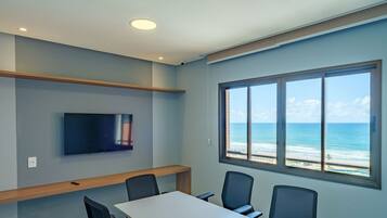 A conference room with a large window overlooking the ocean, a flat-screen TV mounted on the wall, and a wooden shelf above the table.