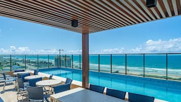 A rooftop infinity pool with a view of the ocean, outdoor seating, and a covered area with a striped ceiling.