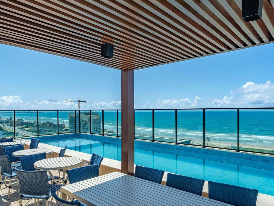 Une piscine à débordement située sur le toit, offrant une vue sur l’océan, ainsi qu’un espace extérieur aménagé et une zone couverte avec un plafond à rayures.