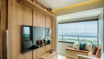 A modern living room with a large flat-screen TV mounted on a wooden wall, a seating area with cushions, and a large window offering a view of the ocean.