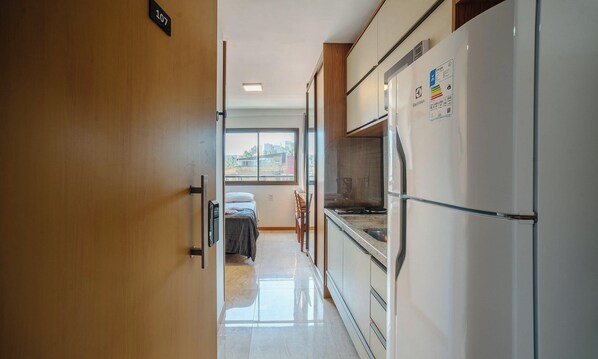 A hotel room with a wooden door, a bed, a small table, and a kitchenette with a refrigerator.