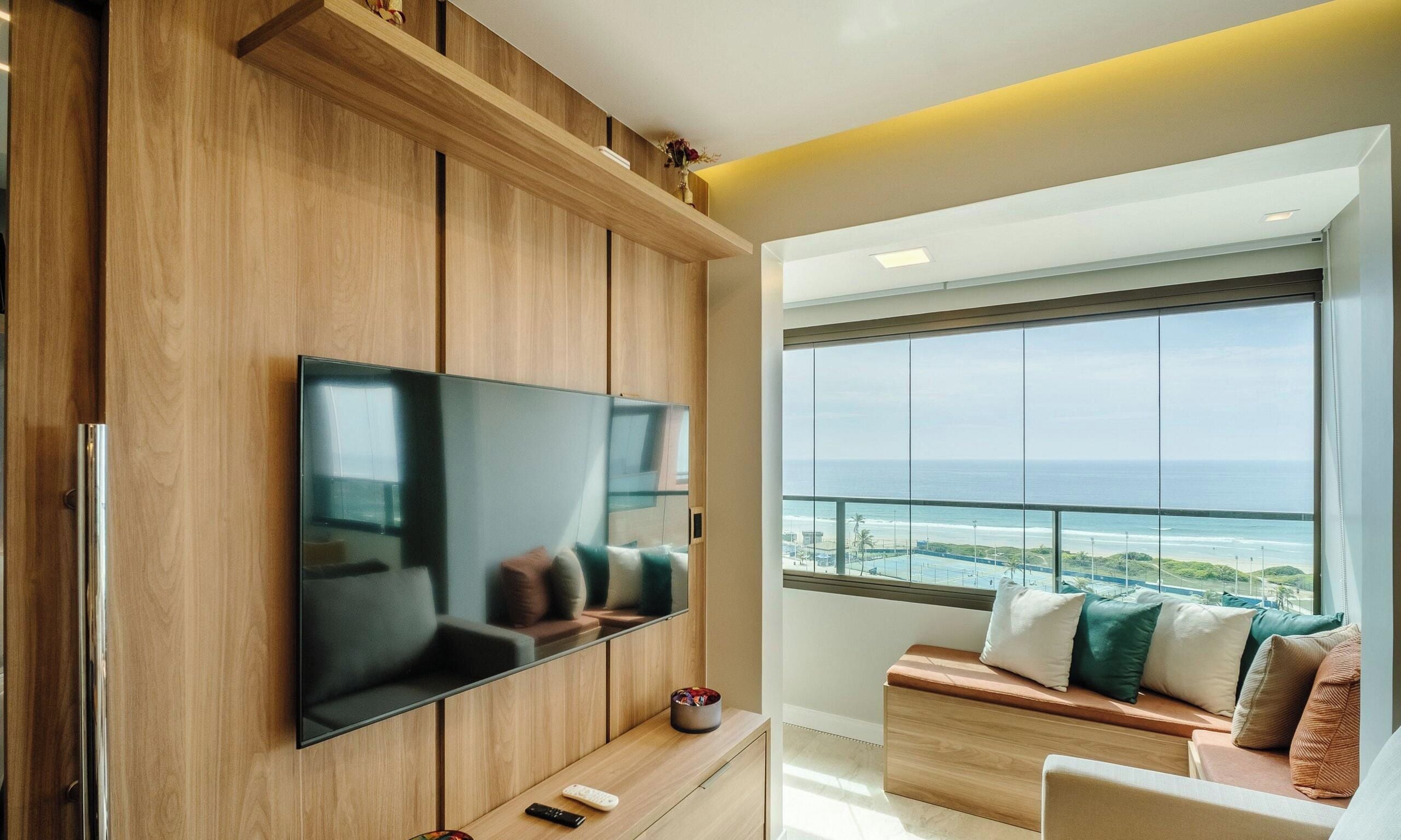 A modern living room with a flat-screen TV mounted on a wooden wall, a sofa with cushions, a small table with a remote control, and a large window offering a view of the ocean.