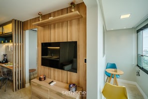 A modern living room with a flat-screen TV mounted on a wooden wall, a dining area with a small table and chairs, and a window with a city view.