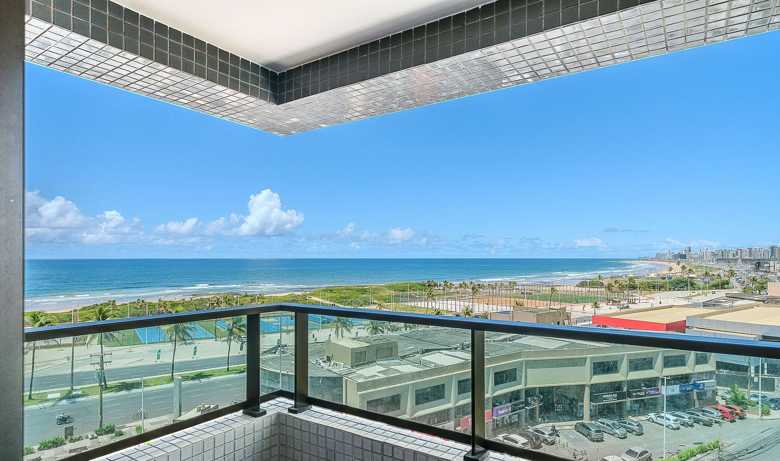 A balcony with a clear view of the beach and ocean, a parking area, and a cityscape in the distance.