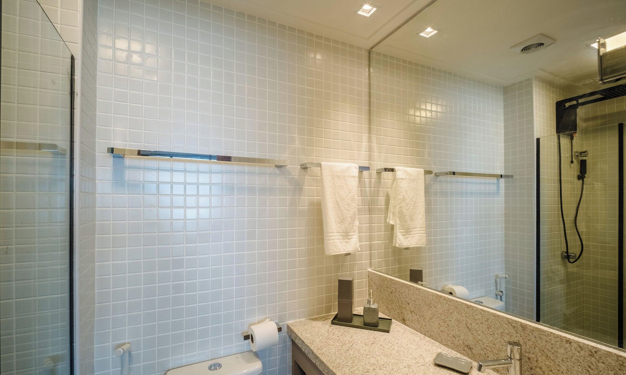 A bathroom with white tiled walls, a glass shower enclosure, a marble countertop, and a toilet with a paper towel dispenser.