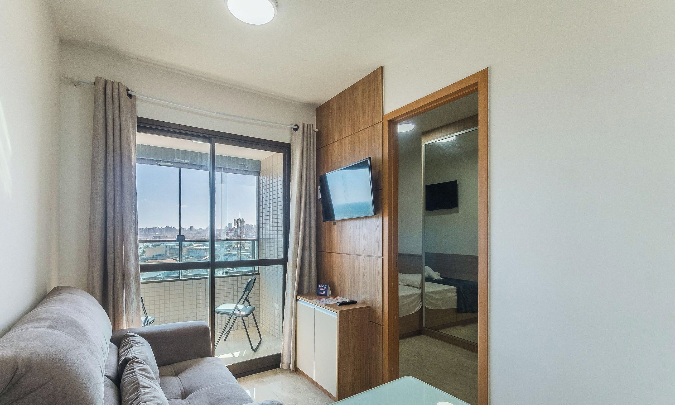 Un salón moderno con sofá, televisión montada en un panel de madera y un balcón con vista a la ciudad.