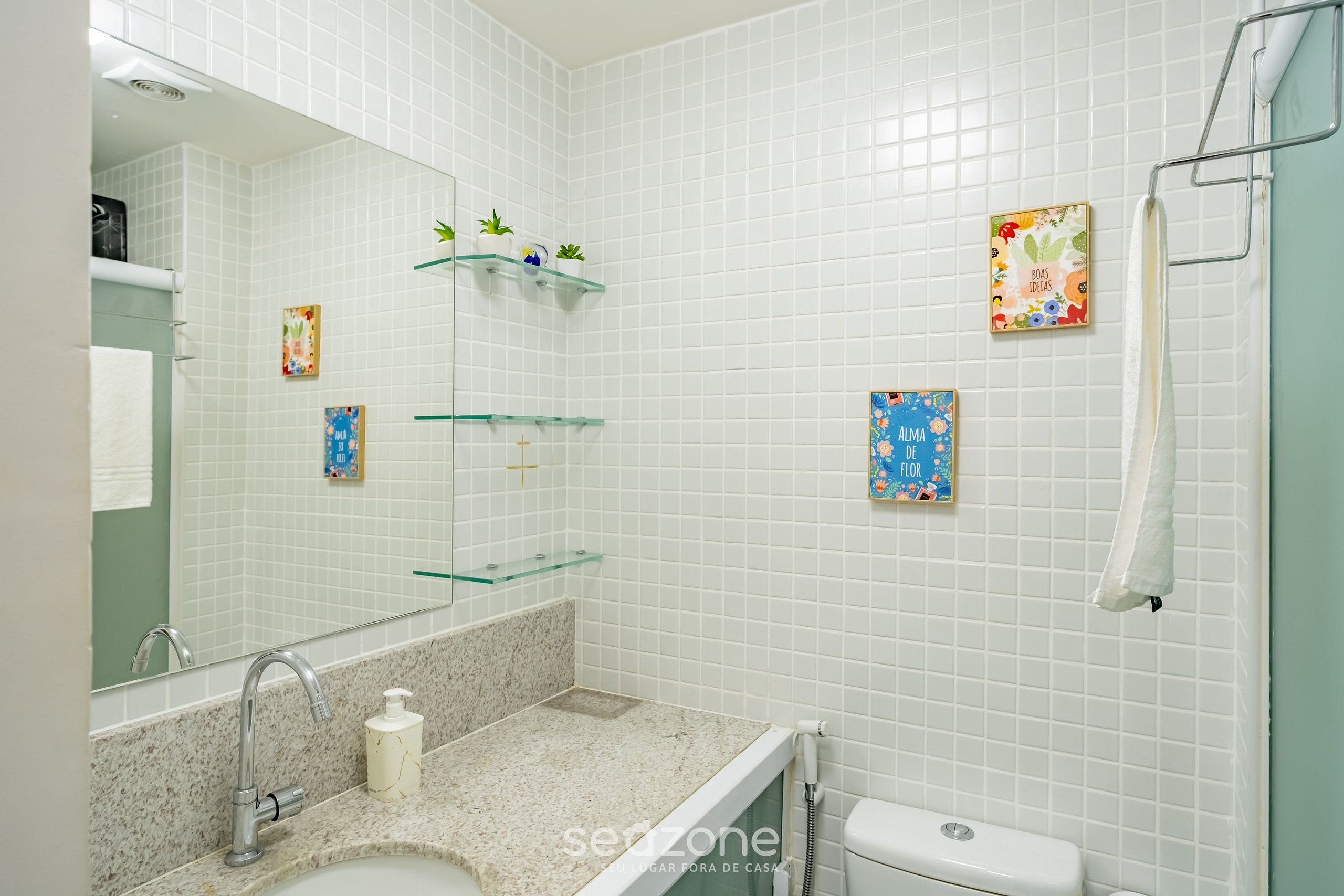A bathroom with white tiled walls, a granite countertop, a sink, a mirror, and a shower area.