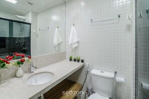 Un baño moderno con lavamanos, espejo y inodoro blancos. Hay dos pequeñas macetas con plantas sobre el mostrador y un box de ducha de vidrio.