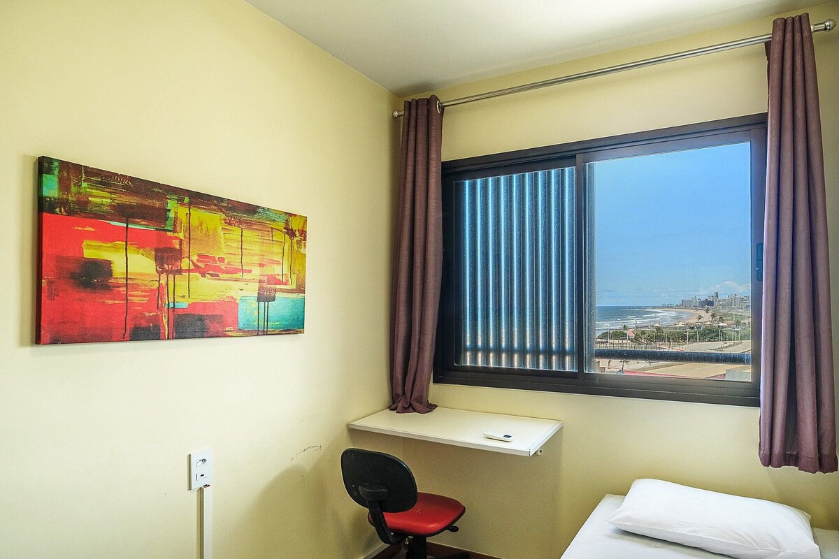 Habitación de hotel con cama, escritorio y silla, ventana con cortinas y un cuadro abstracto de colores en la pared.