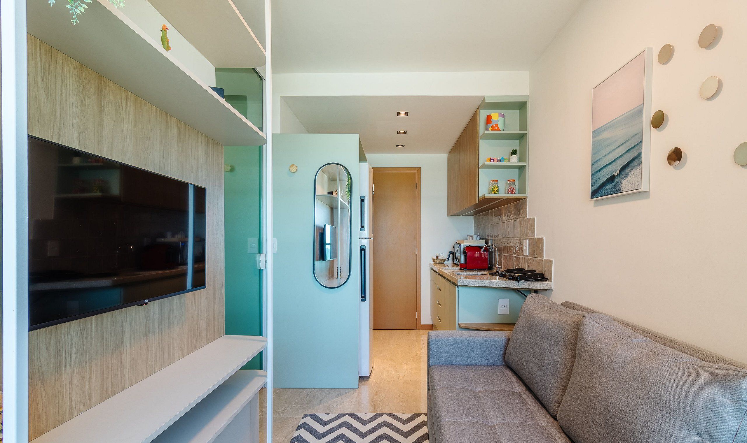 A compact living space with a TV mounted on a wall, a refrigerator, a kitchenette with a stove, and a sofa.