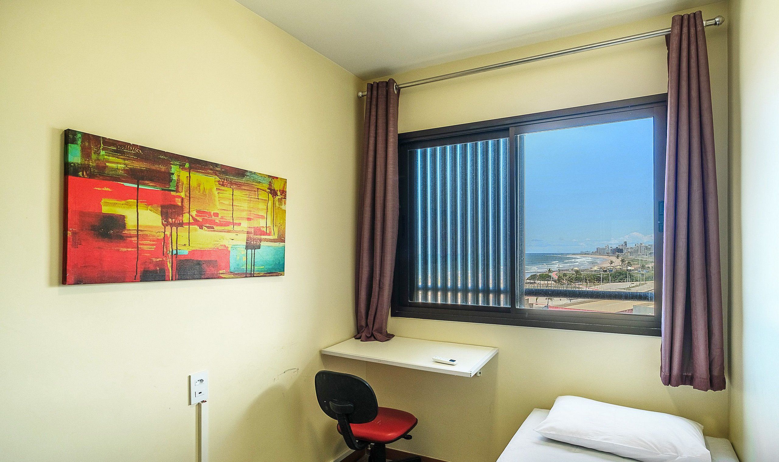 Une chambre d’hôtel avec un lit, un bureau et une chaise, une fenêtre avec des rideaux, et une peinture abstraite aux couleurs vives accrochée au mur.