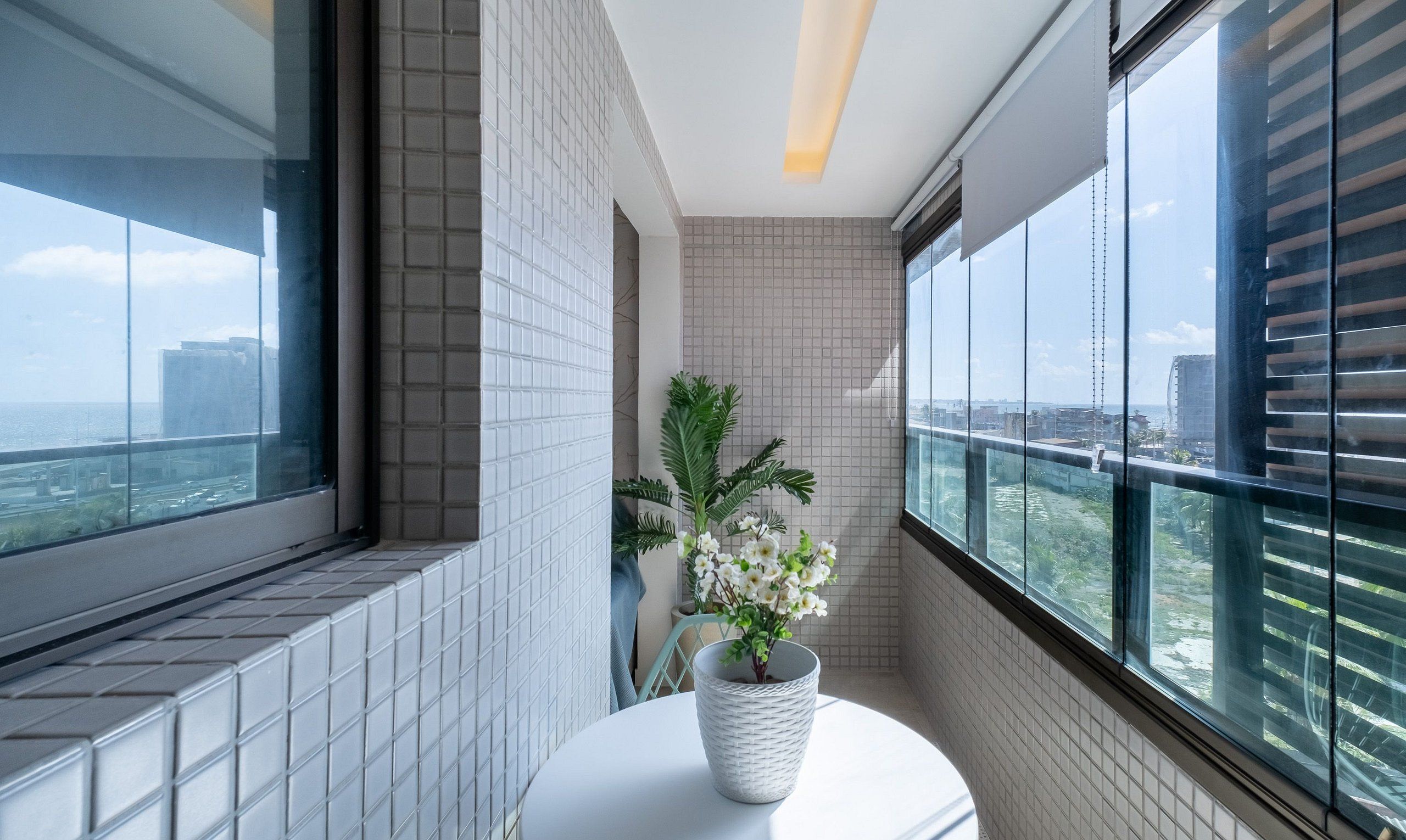 Um banheiro moderno com vista para a cidade através de grandes janelas, parede com azulejos brancos e uma planta em vaso sobre um pedestal branco.