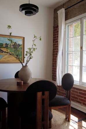 A dining area with a wooden table, chairs, a vase with branches, a painting on the wall, and a window with curtains.