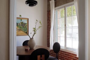 A dining area with a brick wall, a large window with white curtains, a painting on the wall, and a vase with branches on the table.
