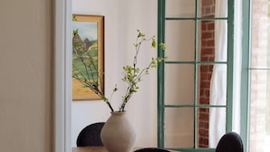 Ein Essbereich mit einem Holztisch, zwei Stühlen, einer Vase mit Zweigen, einem gerahmten Bild und einem grün umrandeten Fenster.