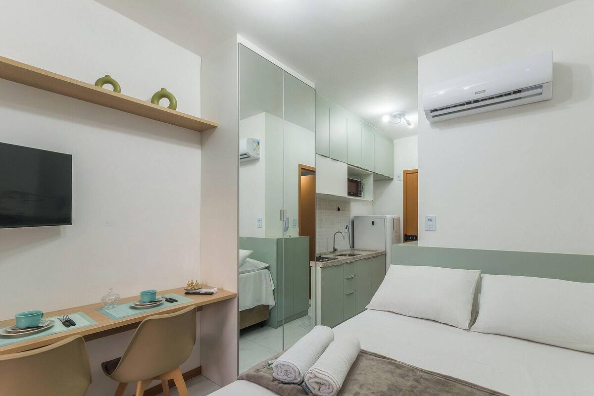 A hotel room with a bed, a desk with a chair, a television, an air conditioning unit, and a bathroom visible through a glass door.