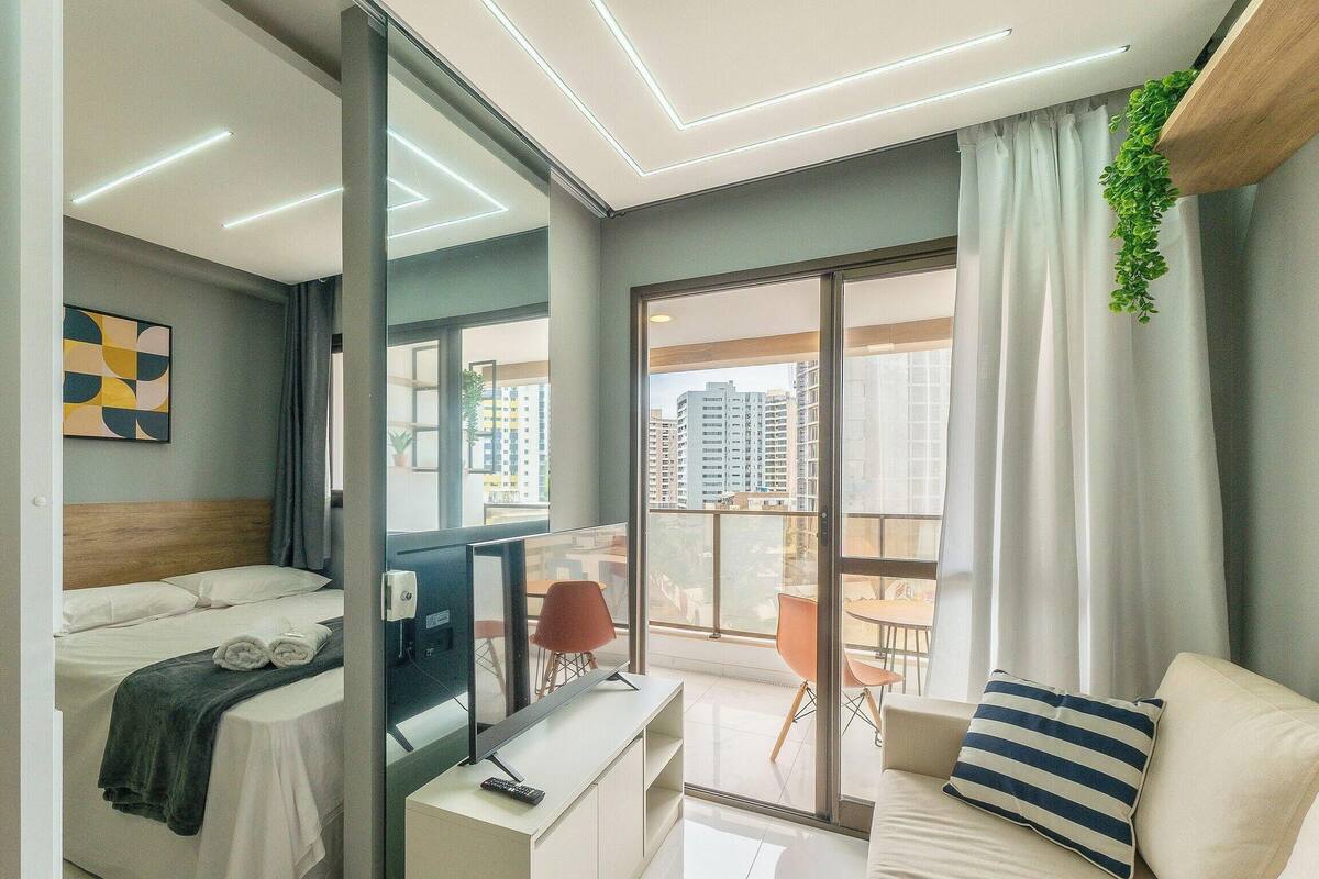 A modern hotel room with a bed, a desk with a computer, a TV, a sofa, and a balcony with a city view.