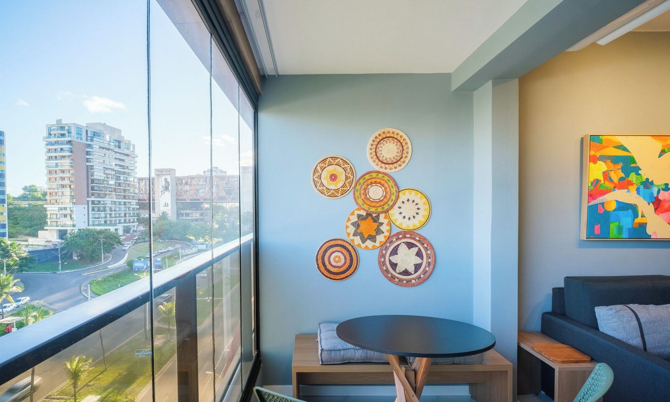 A balcony with a view of a cityscape, a round table with a cushion, and a wall with circular woven decorations.
