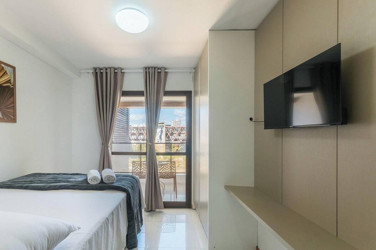 A hotel room with a bed, a flat-screen TV mounted on the wall, a balcony with curtains, and a city view.