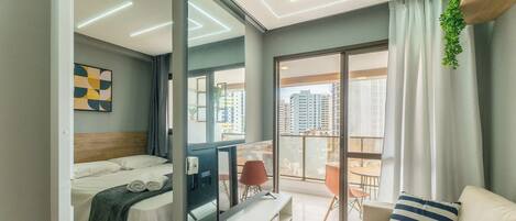 A modern hotel room with a bed, a desk, a chair, a TV, and a balcony with a view of the city.