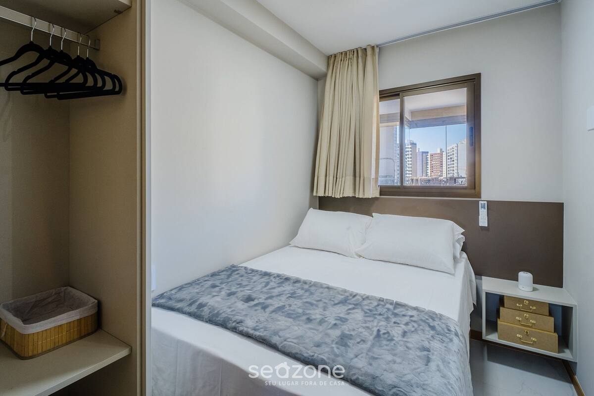 A hotel room with a bed, a window with curtains, a bedside table with drawers, a hanging coat rack, and a small storage box.