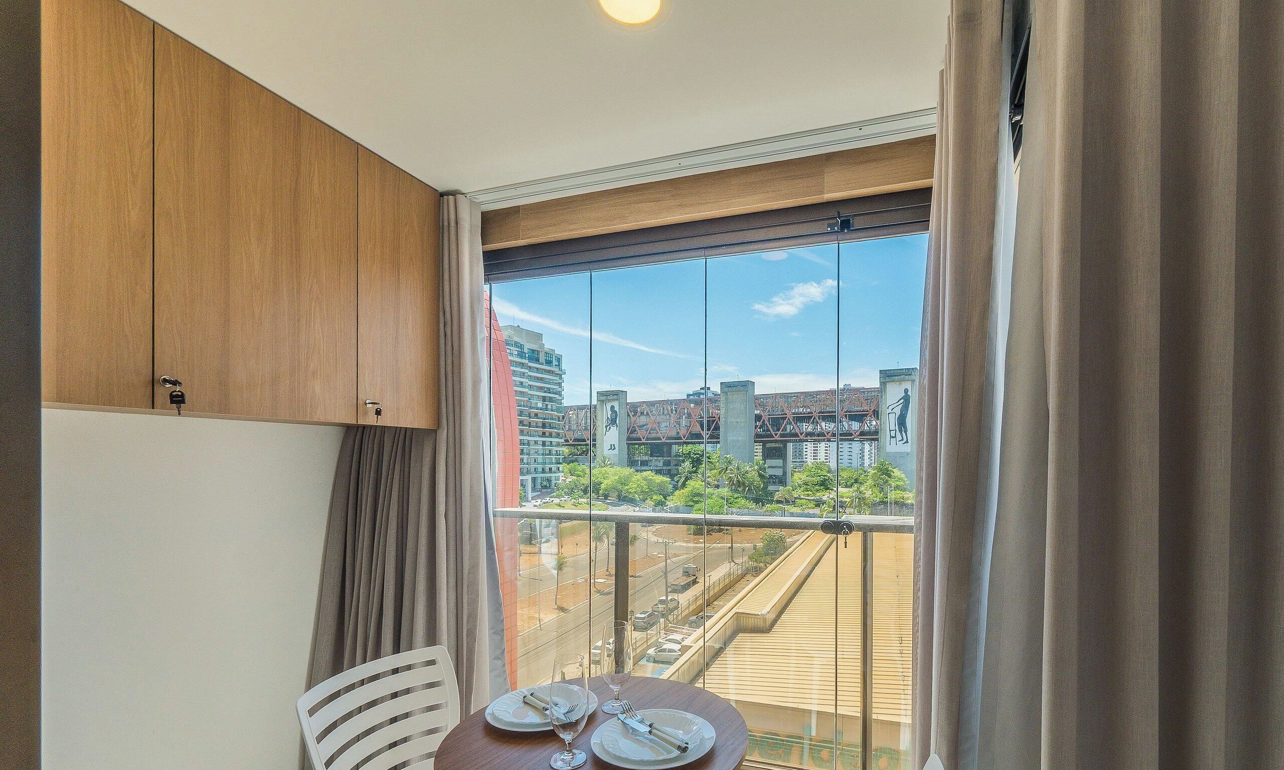 Ein Zimmer mit Esstisch, gedeckt für zwei Personen, Blick auf eine Stadtkulisse durch ein großes Fenster und einer Holzkommode.