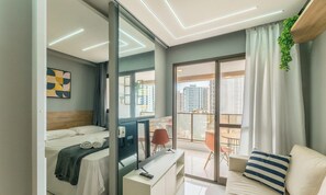 Une chambre d’hôtel moderne équipée d’un lit, d’un bureau avec un ordinateur, d’une télévision, d’un canapé et d’un balcon offrant une vue sur la ville.