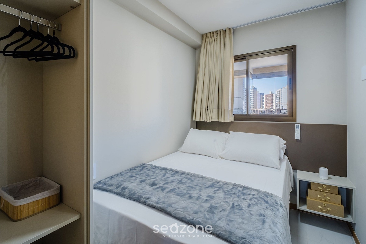 Une chambre d’hôtel comprenant un lit, une fenêtre avec des rideaux, une table de chevet avec des tiroirs, un porte-manteau et une petite boîte de rangement.