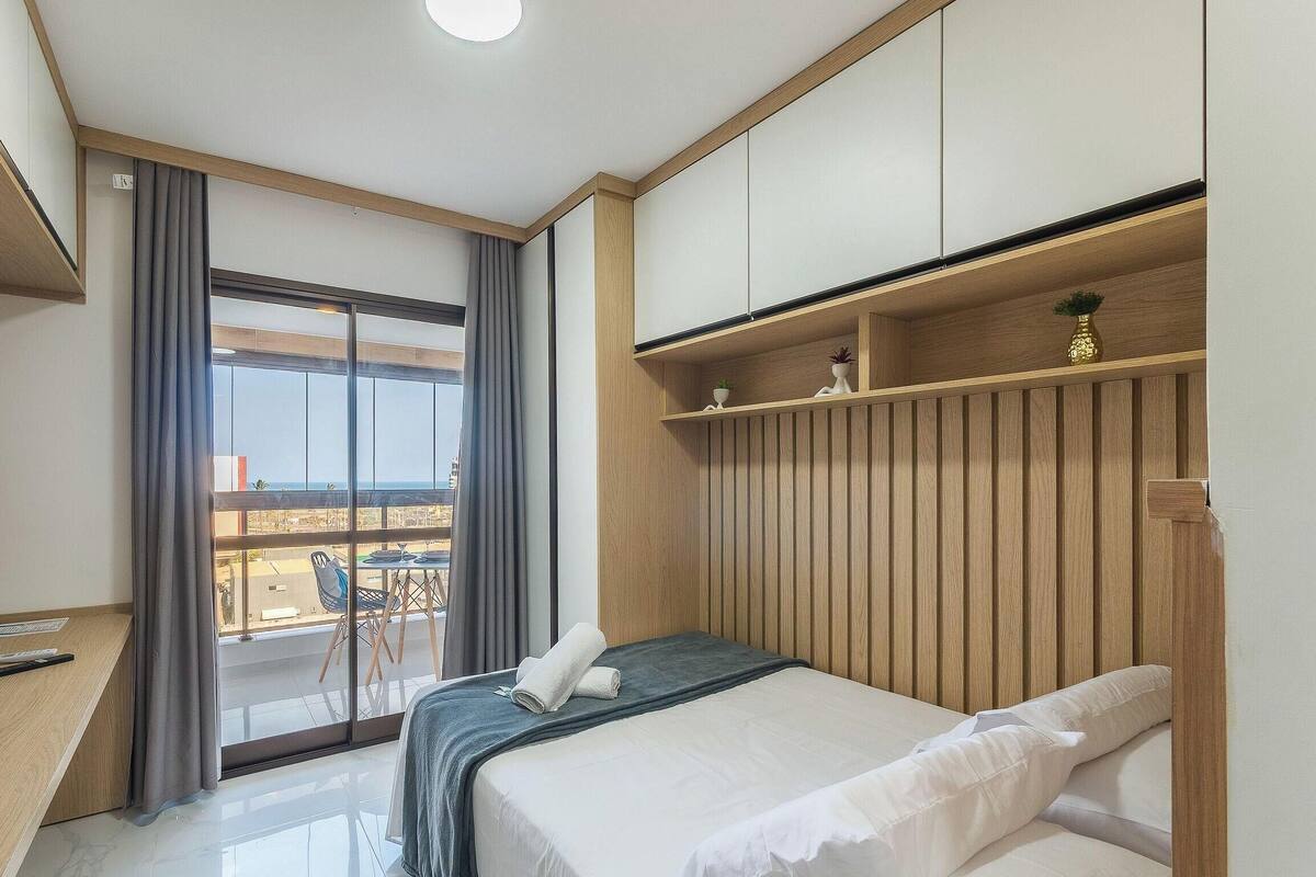 A hotel room with a bed, a desk, a chair, a balcony with a view, and a wooden wardrobe.
