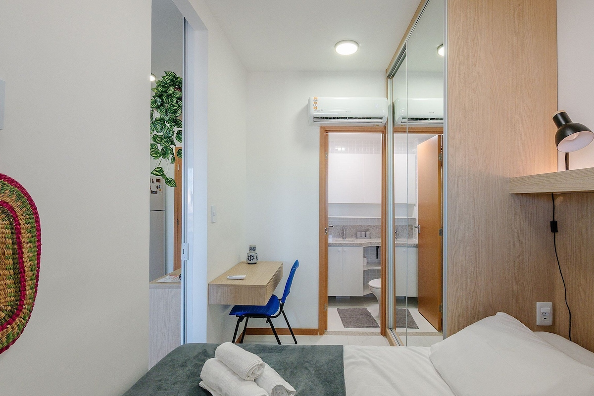 Une chambre d’hôtel de taille réduite, comprenant un lit, un bureau, une chaise, un miroir et un climatiseur fixé au mur.