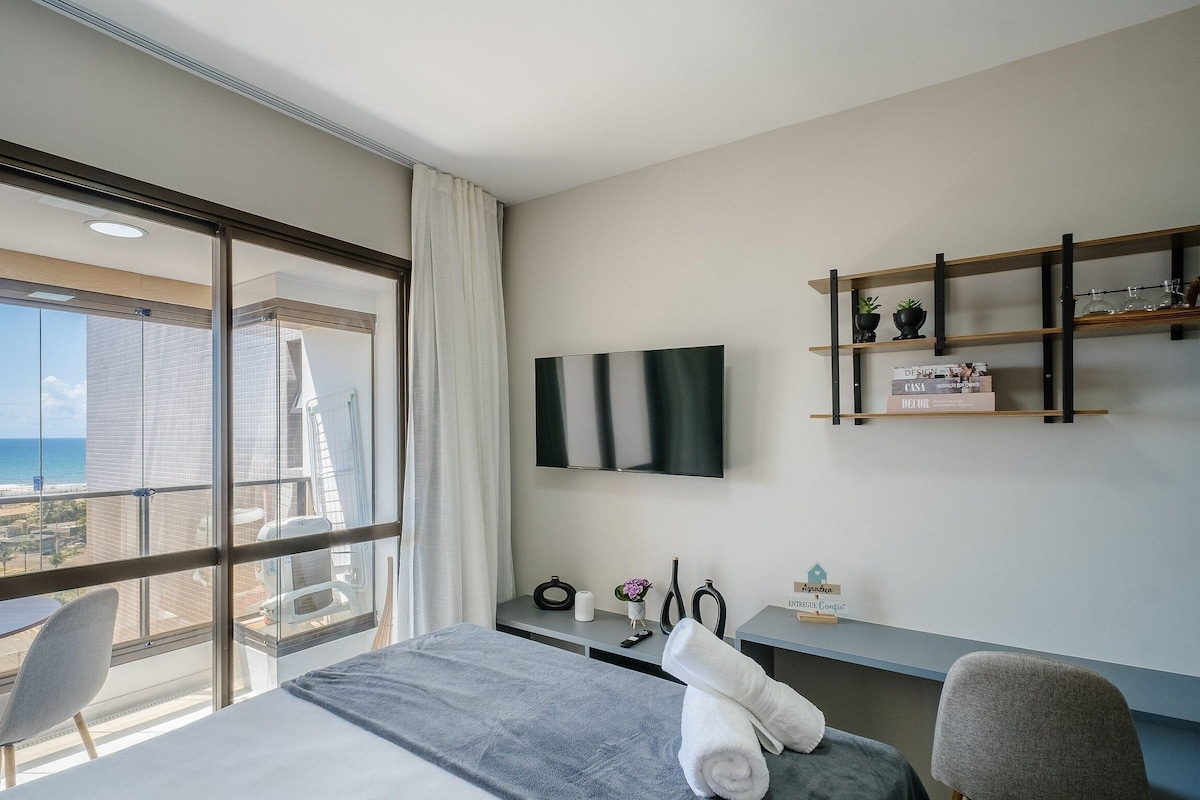 Une chambre d’hôtel dotée d’une grande fenêtre, d’un lit, d’un bureau, d’une chaise, d’un téléviseur fixé au mur et d’une petite étagère ornée d’objets décoratifs.