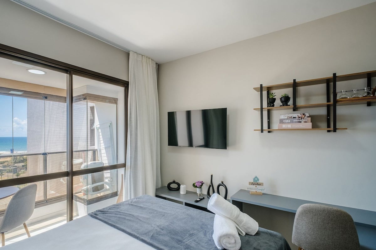 A hotel room with a large window, a bed, a desk, a chair, a TV mounted on the wall, and a small shelf with decorative items.