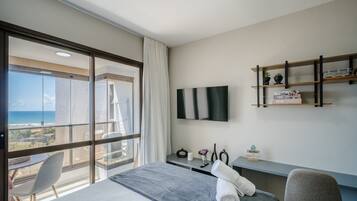 Habitación de hotel con un ventanal grande, una cama, un escritorio, una silla, un televisor montado en la pared y un pequeño estante con objetos decorativos.
