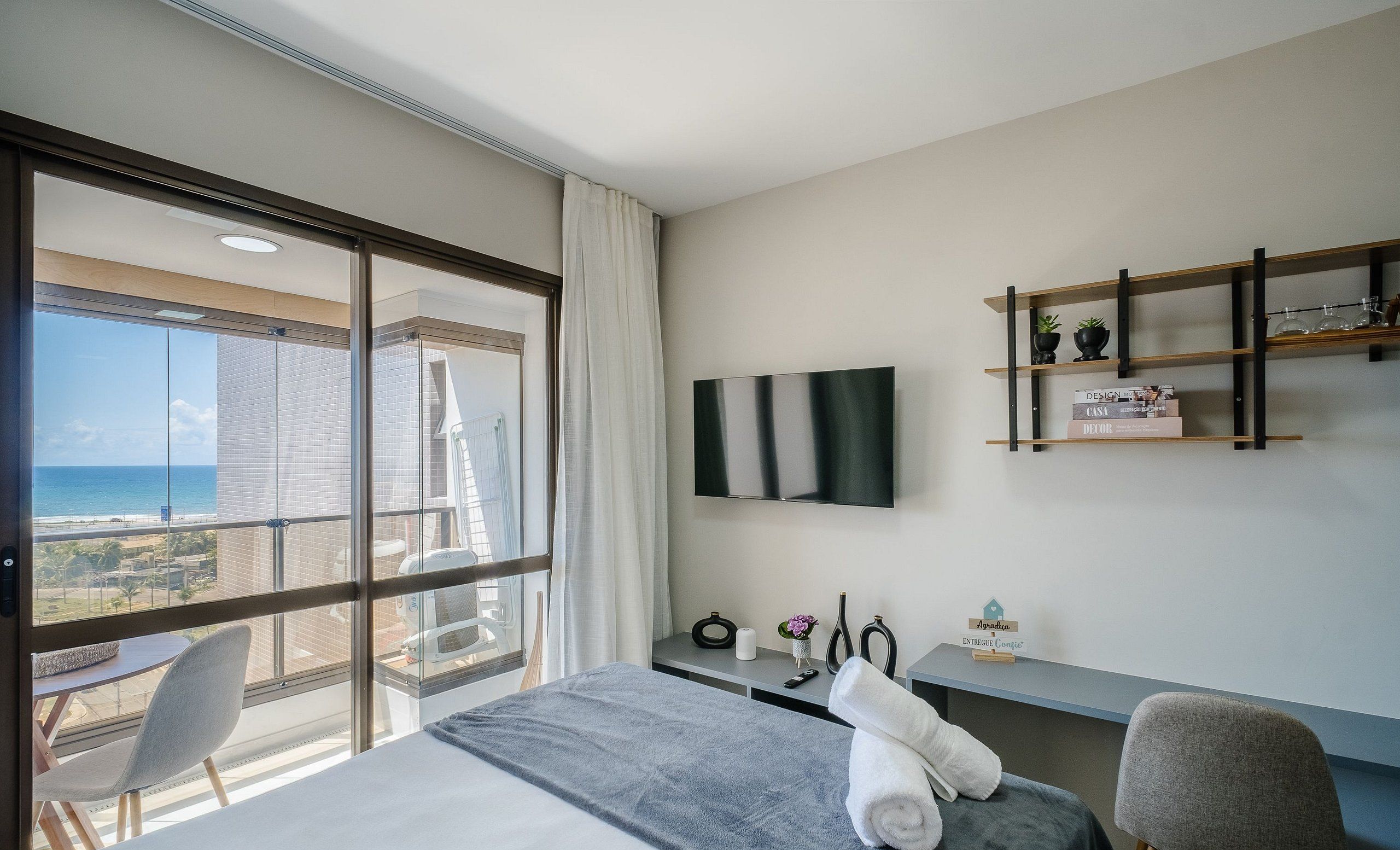 A hotel room with a large window, a bed, a desk, a chair, a TV mounted on the wall, and a small shelf with decorative items.