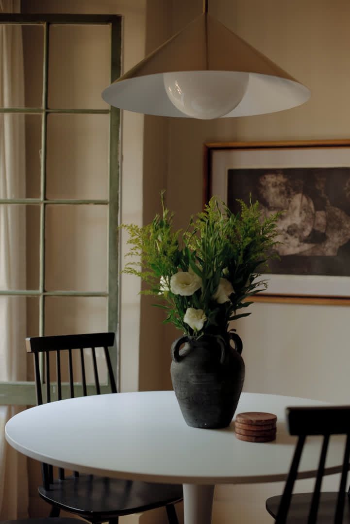 A round table with a vase of flowers, two chairs, a pendant light, and a framed picture on the wall.