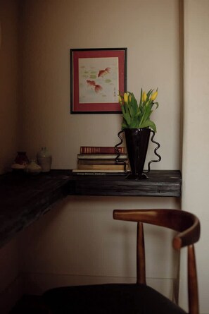 Een hoek van een kamer met een houten stoel, een zwarte vaas met gele bloemen, een lijst met een afbeelding aan de muur en een stapel boeken op een plank.
