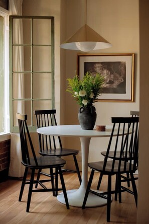A dining area with a white round table, four black chairs, a vase with flowers, a framed picture on the wall, and a window with a curtain. - Elegant Studio in Hollywood (Los Angeles)