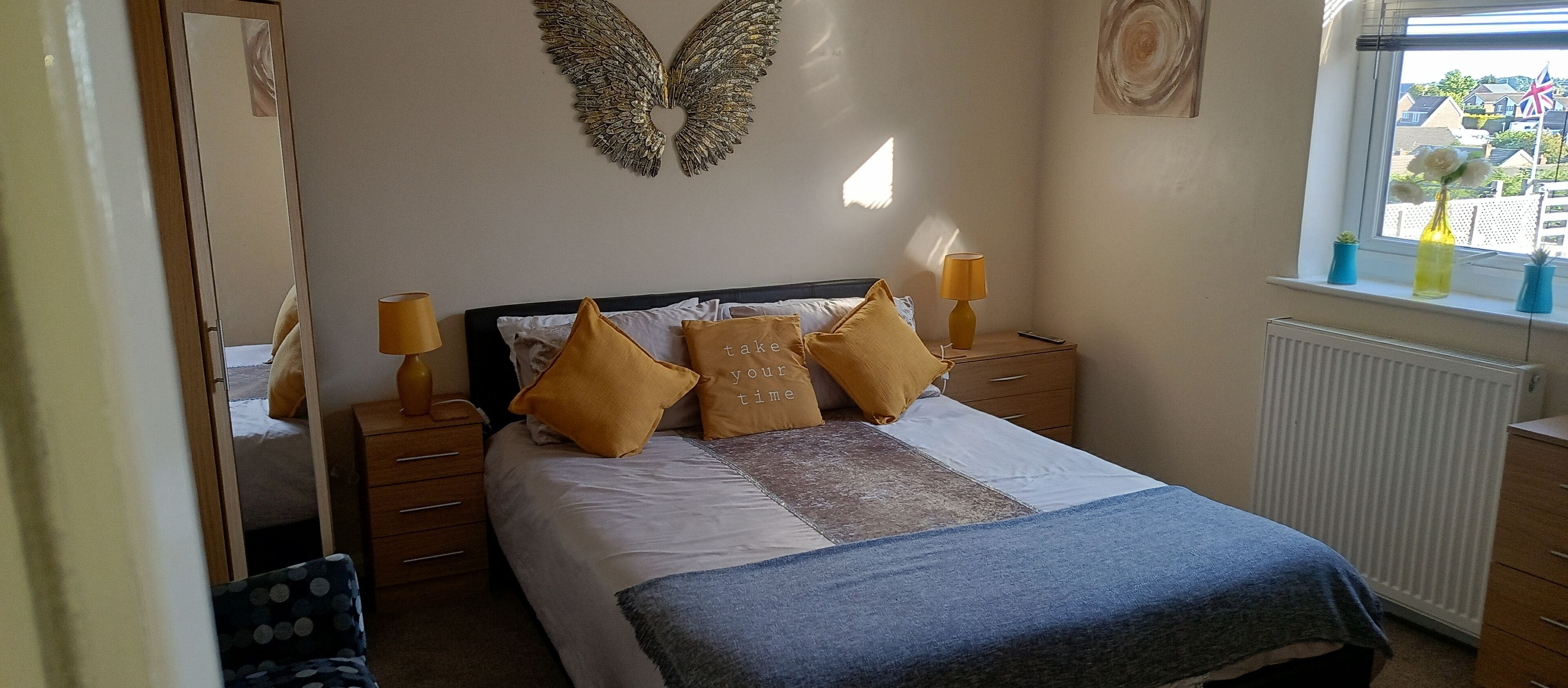 A bedroom with a bed, a nightstand with lamps, a dresser, a radiator, and a window with a view of houses and a flag.