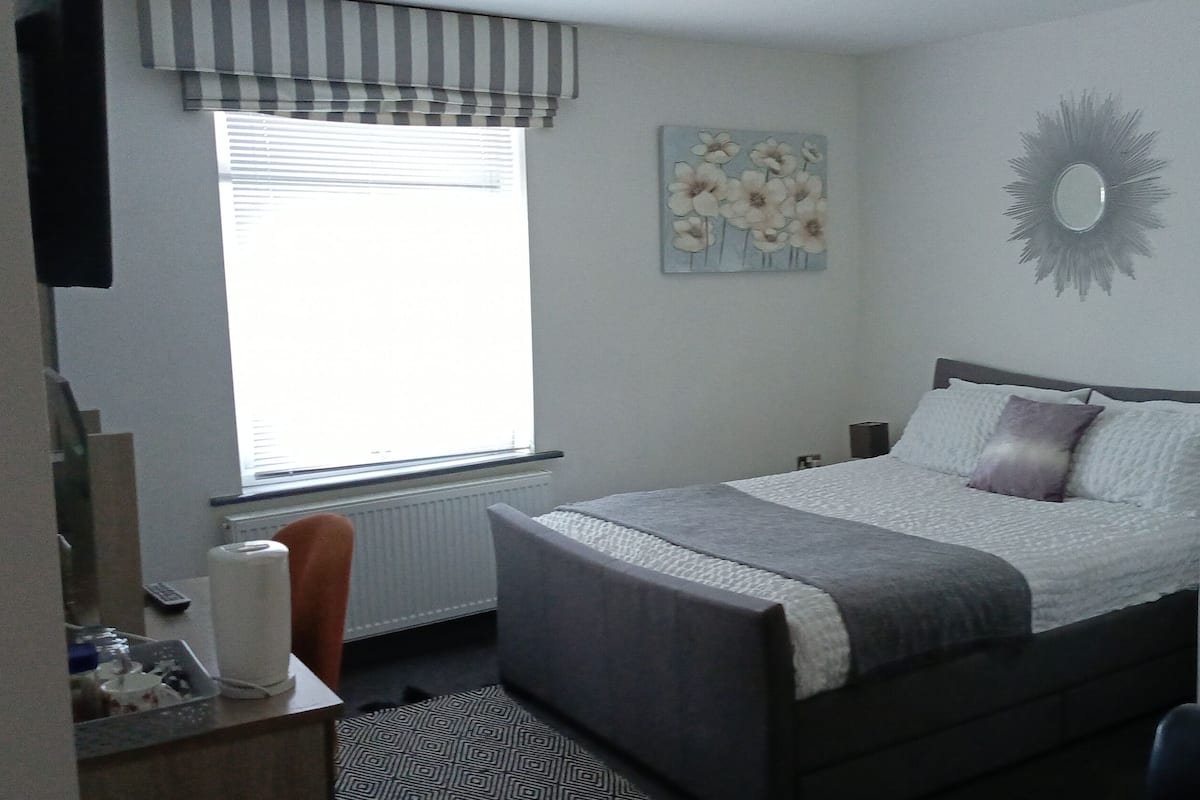 A bedroom with a bed, a window with blinds, a radiator, a bedside table with a lamp, and a wall decoration.