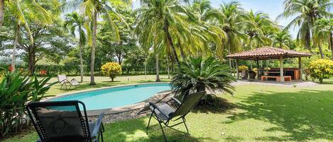 A poolside area with lounge chairs, a covered seating area, and lush greenery.