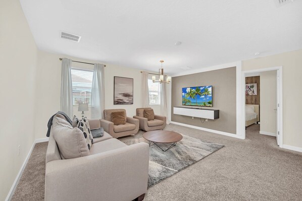 A living room with a sofa, two armchairs, a coffee table, a TV mounted on the wall, and a door leading to a bedroom.
