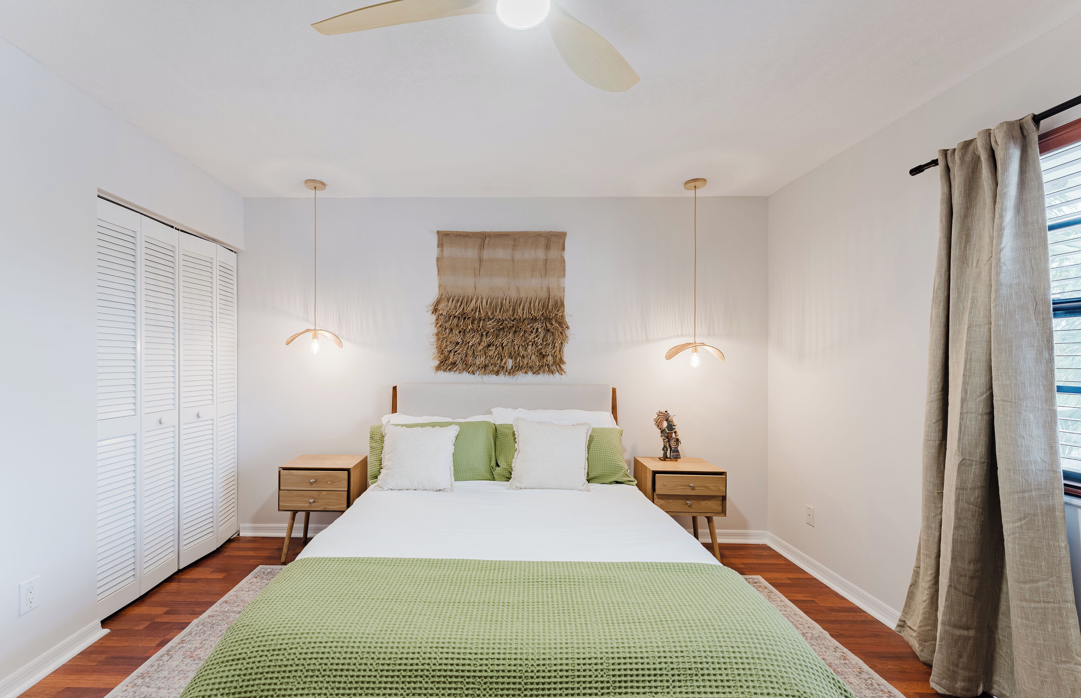 A bedroom with a bed, bedside tables, a ceiling fan, a wall-mounted light fixture, and a window with curtains.