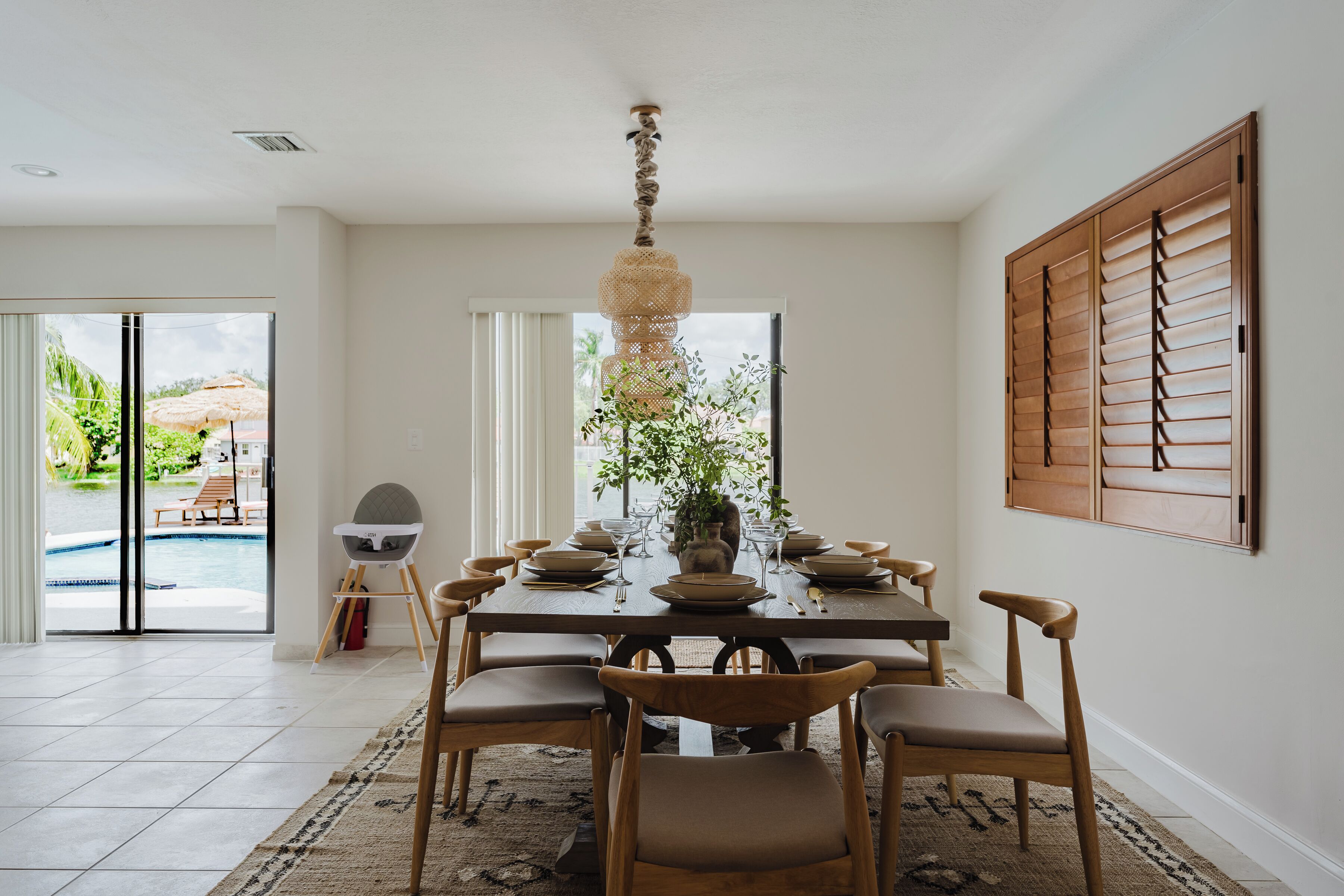 Un coin repas avec une grande table dressée pour six personnes, entourée de chaises en bois, d’un luminaire suspendu et offrant une vue sur une piscine à travers des portes vitrées.