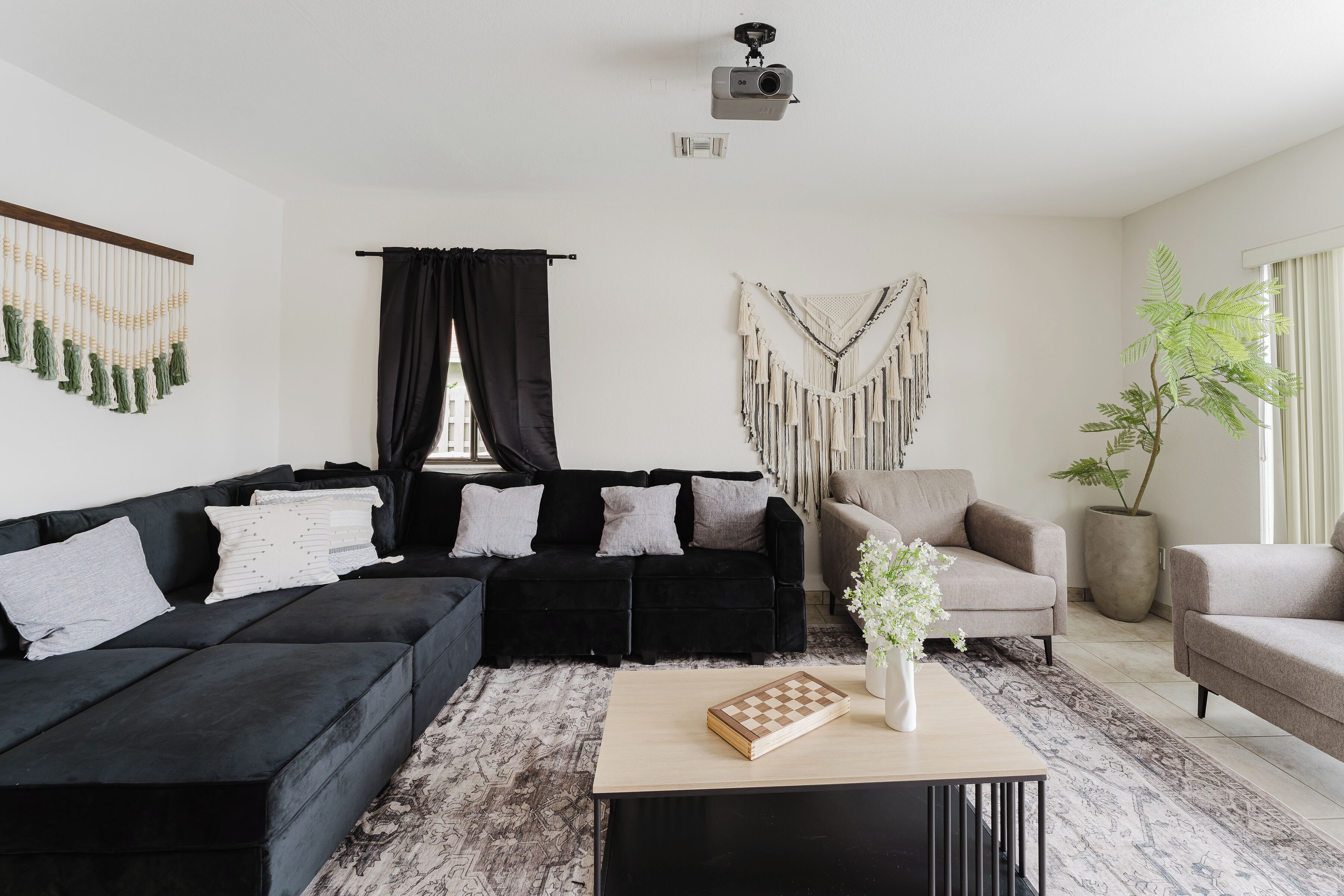 Un salon moderne avec un canapé d’angle noir, un fauteuil beige, une petite table basse surmontée d’un jeu d’échecs, une décoration murale en macramé et une plante en pot.